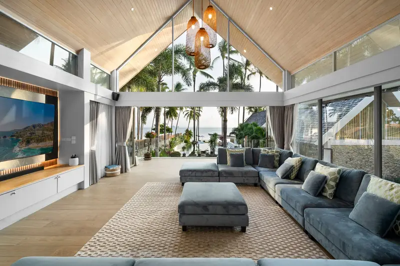 Airy lounge with blue sofas and a high timber ceiling, opening towards palms and the pool.