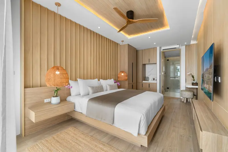 Bedroom 3: King bed with a timber headboard wall, a ceiling fan in a lit coffered ceiling, a wall-mounted television and the en-suite bathroom seen through the open doorway.