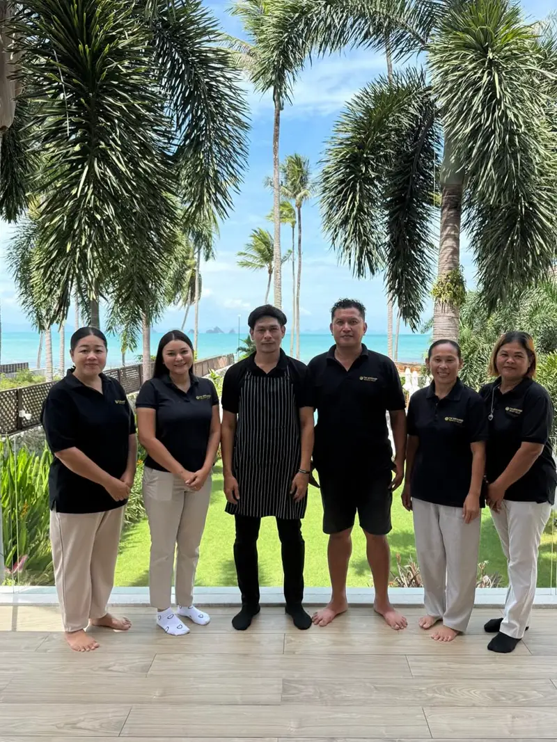 Six members of the Mandarin Beach Villa team, the chef in a striped apron, standing together on the beachfront terrace with palms, the lawn and the sea behind them.