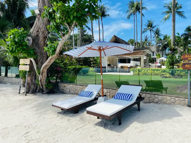 Two loungers with an umbrella beneath a tree on the lawn beside the beach.
