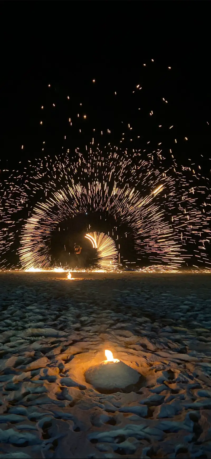 A performer spins fire on the beach at night, sparks arcing over the sand.
