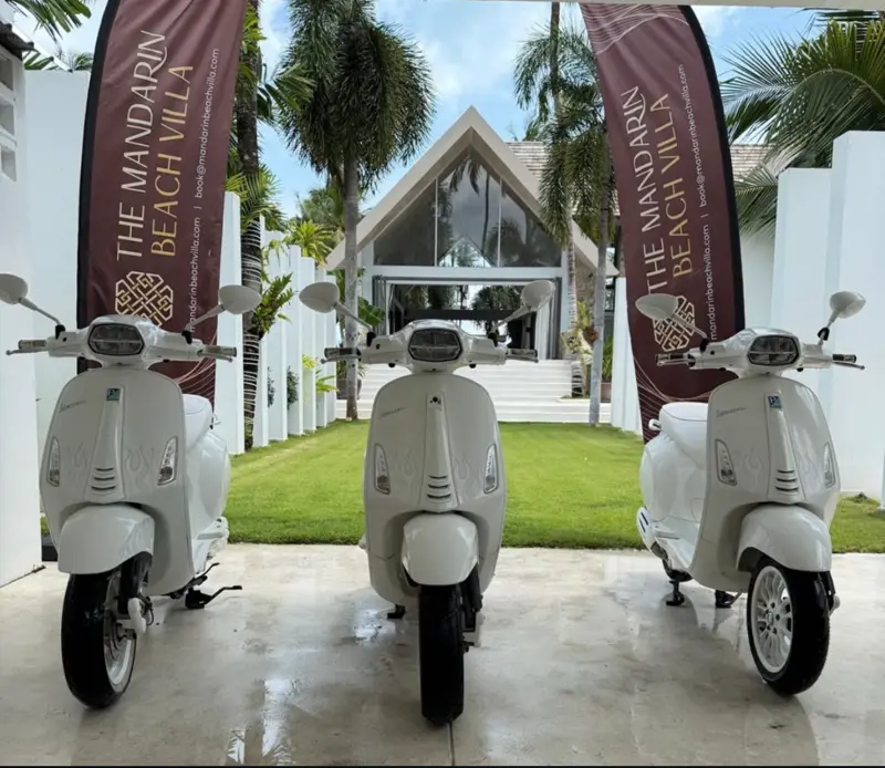Three white Vespa scooters lined up at the entrance of Mandarin Beach Villa, with the villa’s banners either side and the glass gable of the main house behind.