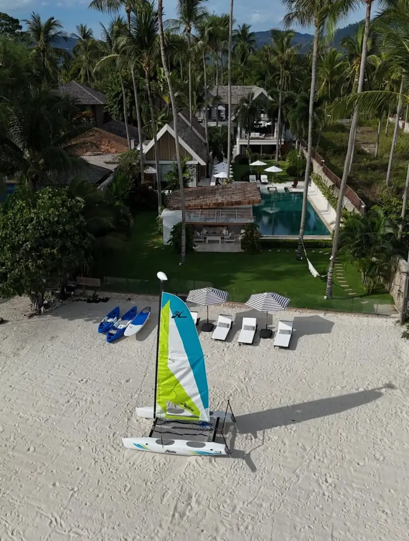 Aerial view of Mandarin Beach Villa’s beachfront: the Hobie Cat and kayaks on the sand, striped umbrellas and loungers, the lawn, the sala and the pool, with the main house behind.
