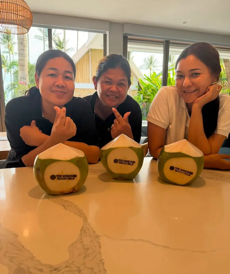 Three members of the Mandarin Beach Villa team smiling at the dining table behind three fresh coconuts stamped with the villa’s name.