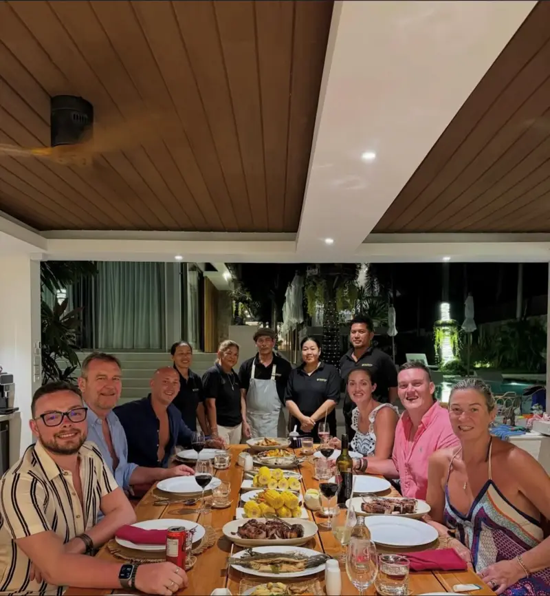 Guests seated at a long dinner table on Mandarin Beach Villa’s terrace with dishes of grilled fish, meat and corn, the chef in his apron and four members of the villa team standing behind them.
